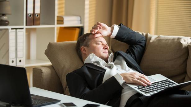 Businessman in suit lying on a couch with two cellphones and laptops, sleeping. Exhausted man relaxing in office early morning. Responsible executive working fell asleep.