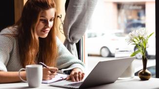 attractive woman writing in notepad while watching webinar on laptop in cafe